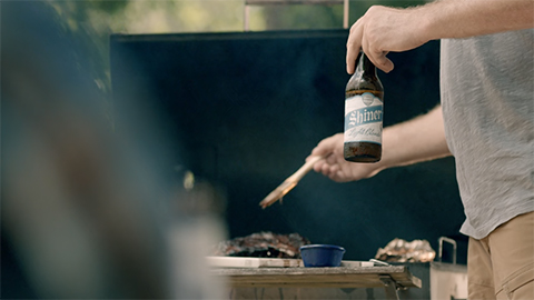 Person holding a Shiner beer while basting meat on a BBQ grill