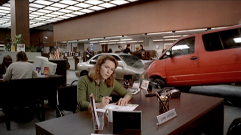 Woman sitting at desk with other people, desks, and cars in the background
