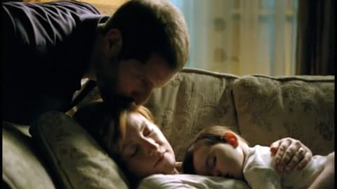 Man kissing woman on the head as she sleeps on couch with baby lying on her chest