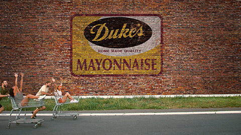 People riding in shopping carts in front of brick wall with Duke's Mayonnaise sign painted on it